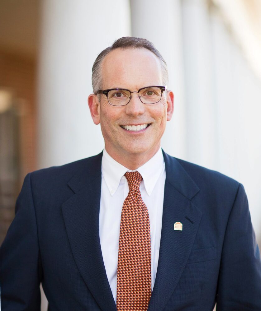 Gary Nimax in a suit and tie smiles confidently in front of a building.