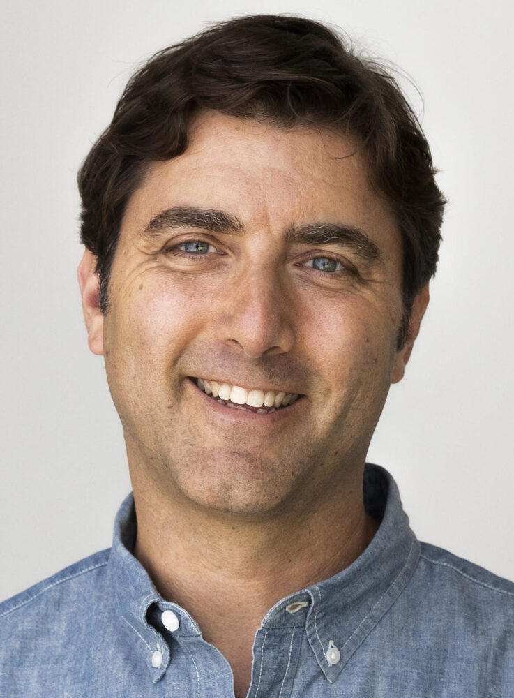 David Koplan portrait. Man with blue eyes smiling in a blue button-down shirt.