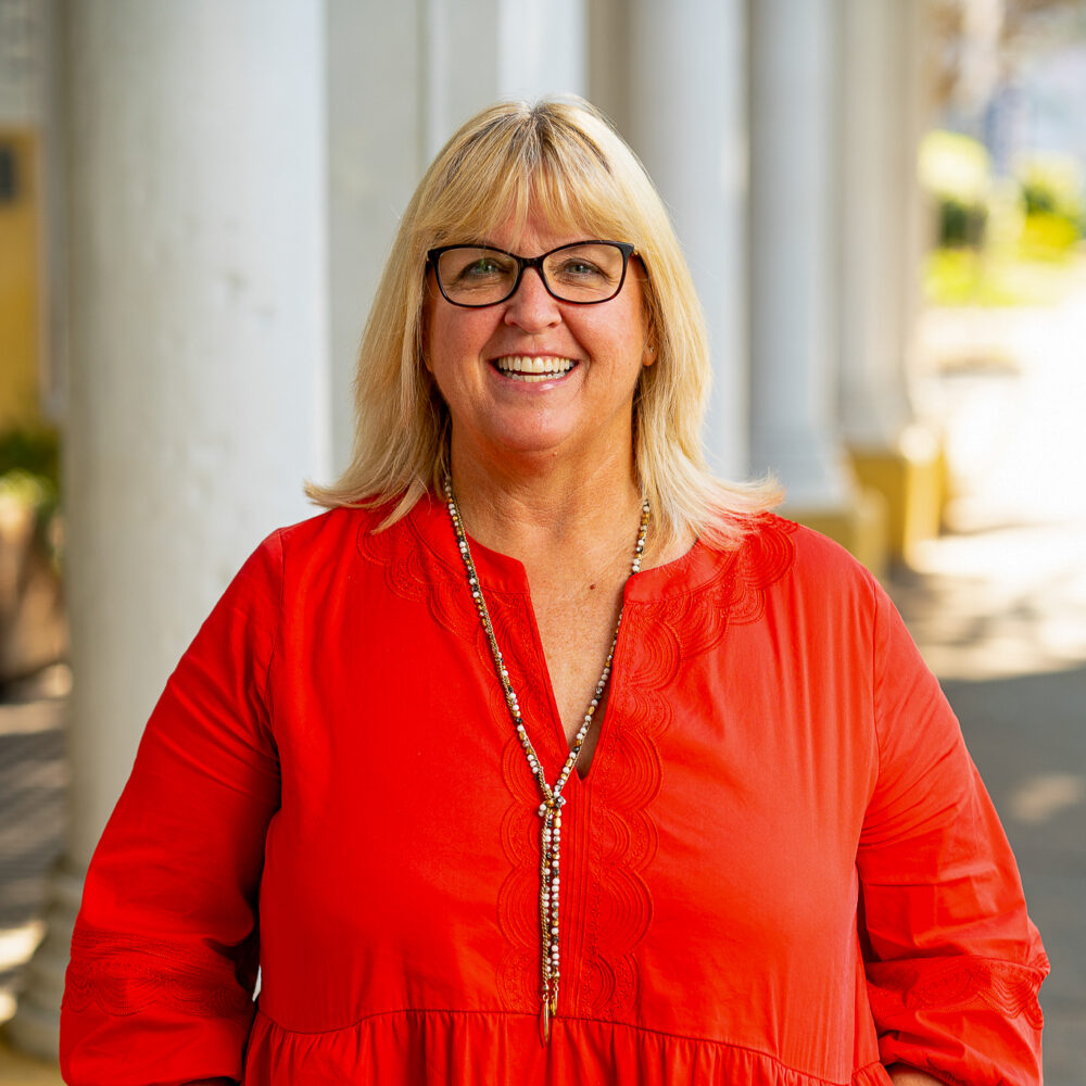 Jenny Gardiner, author, smiles in a red dress with white columns in the background.