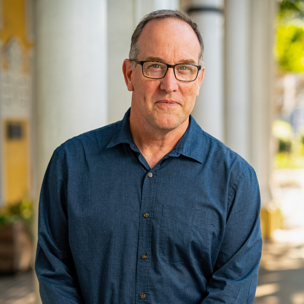 Portrait of John Kelly wearing glasses and a blue shirt.