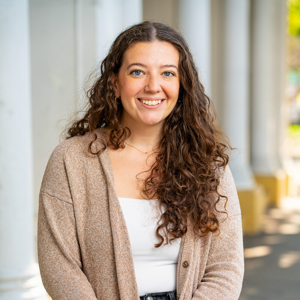 Sarah Ross smiling with curly hair and blue eyes standing outdoors, wearing a cardigan.