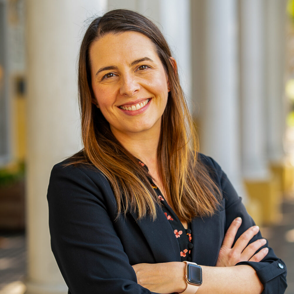 Jenny Mays, smiling woman in a black blazer with arms crossed stands in front of columns.