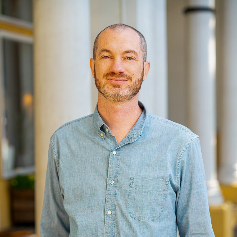 Ilya Tovbis smiles in front of classical columns, wearing a blue button-down shirt.