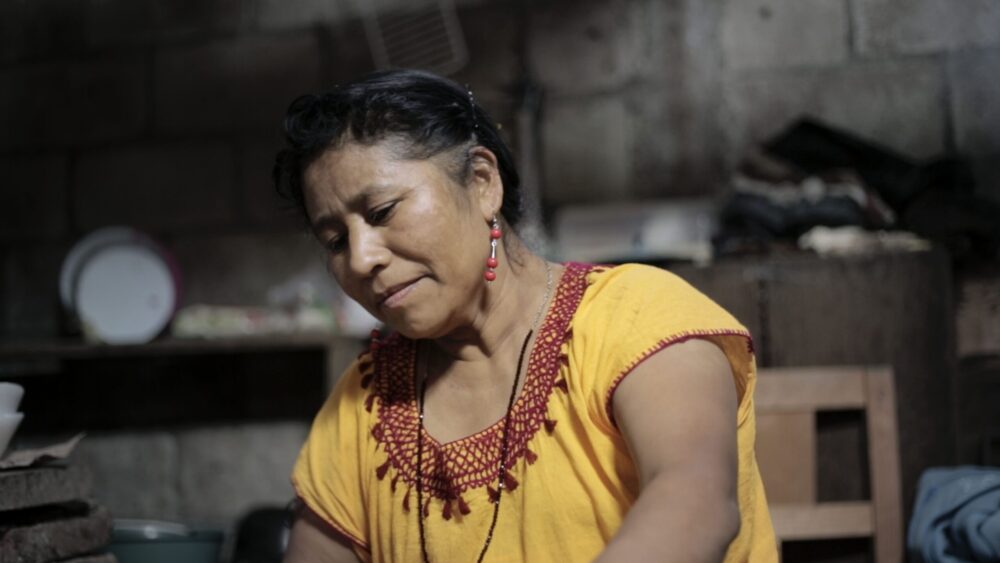 Mom in a yellow blouse, working in a rustic kitchen with stone walls.