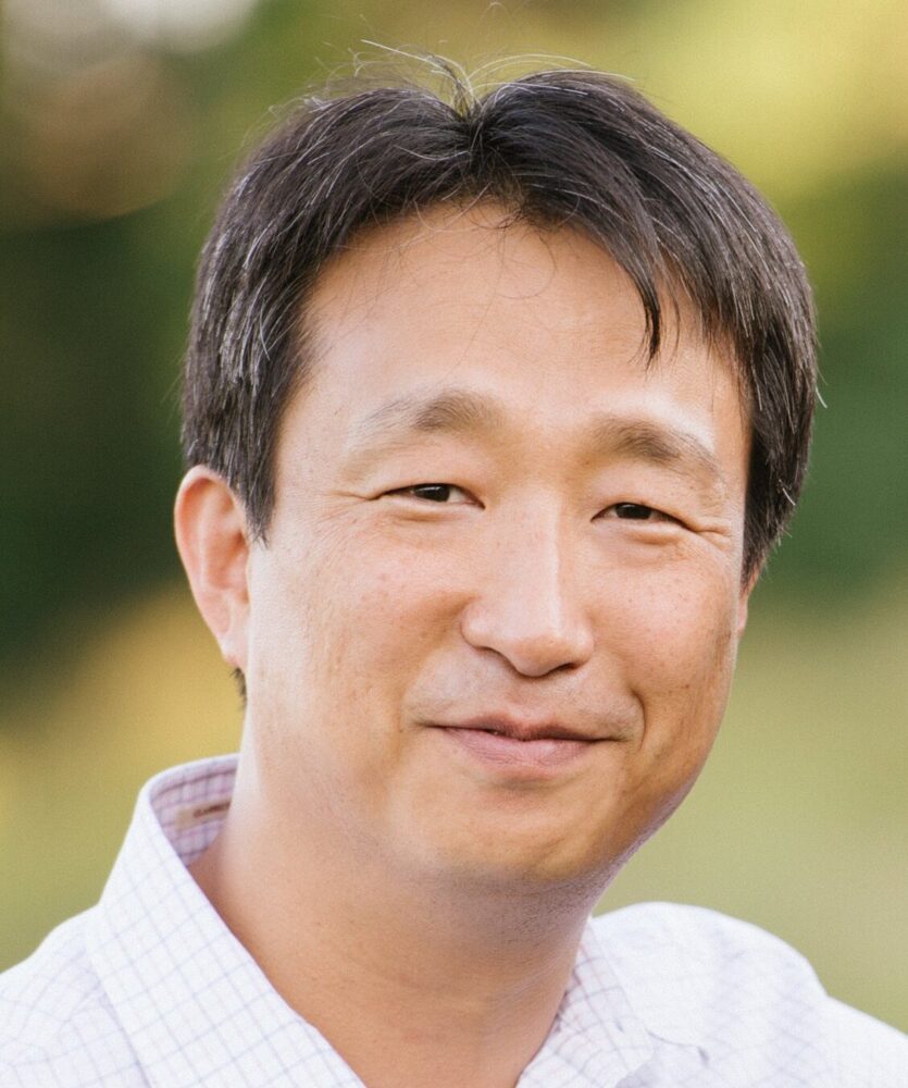 Portrait of Jung-Bum Shin smiling outdoors, wearing a checkered shirt.