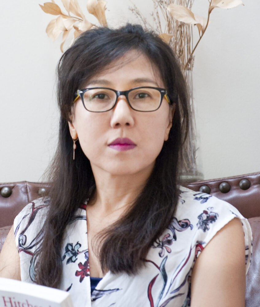 Portrait of Hyeyon Moon wearing glasses, seated on a leather couch with dried flowers in the background.