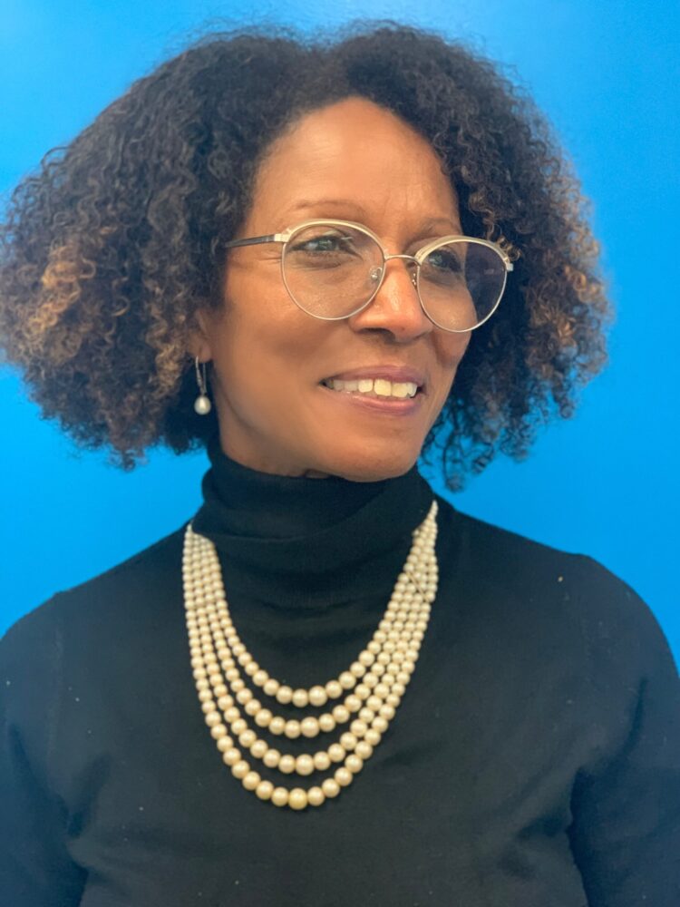 Beverly Adams portrait with pearls, glasses, and a black turtleneck against a blue background.