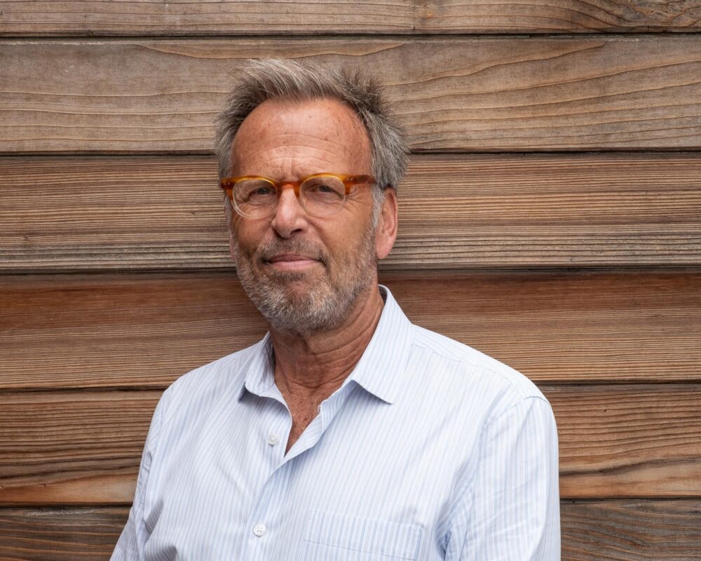 Mark Johnson, Chair, poses in front of a wooden wall, wearing glasses and a light blue shirt.