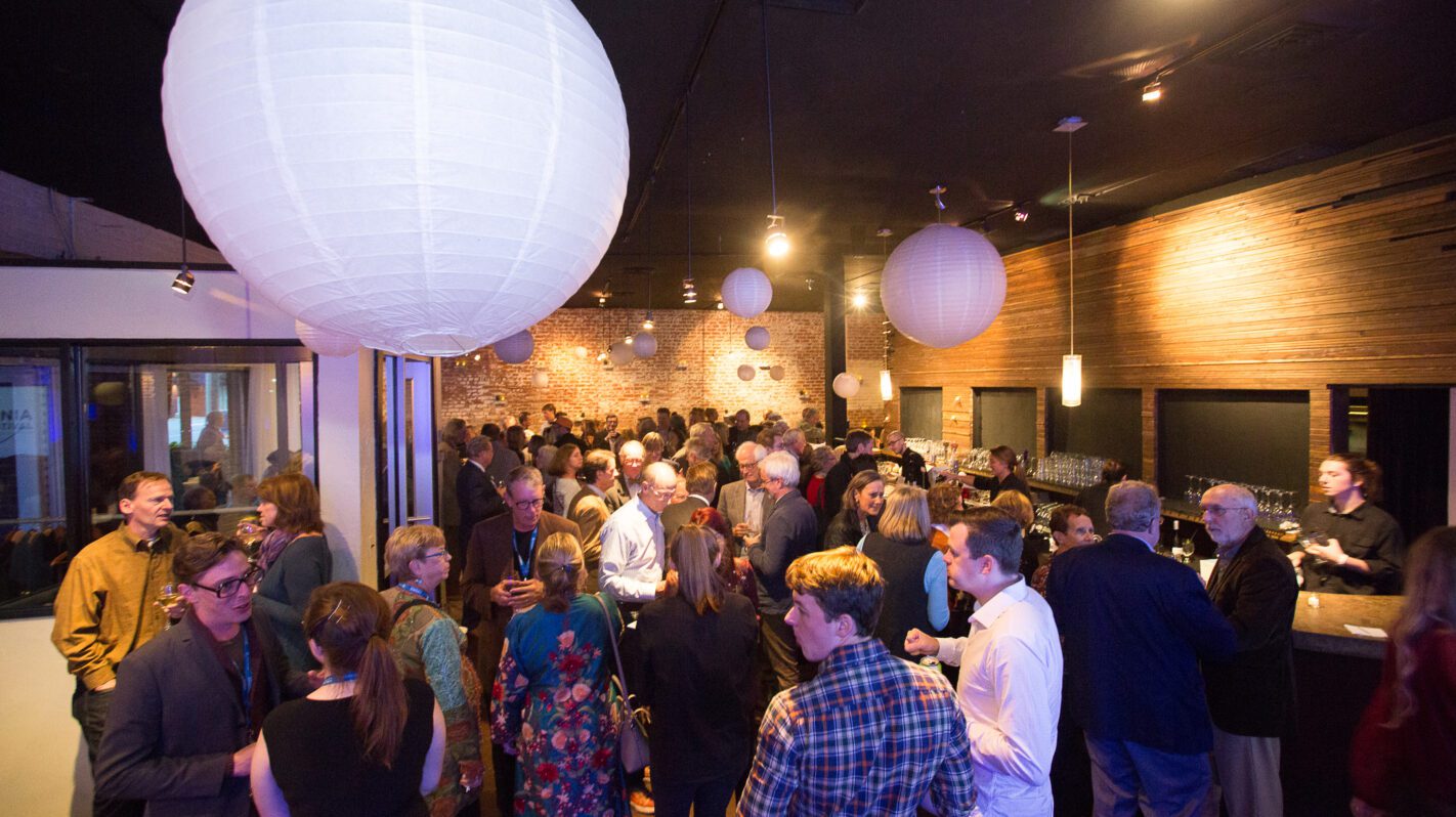 Crowded fundraising event with people mingling and a bar area.