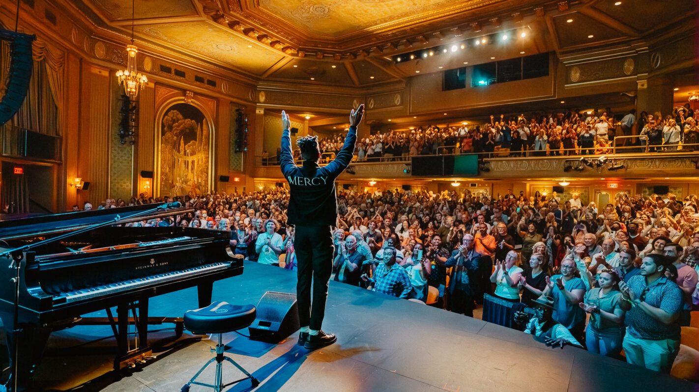 Concert hall with performer on stage, arms raised, audience clapping. "Mercy" on jacket.