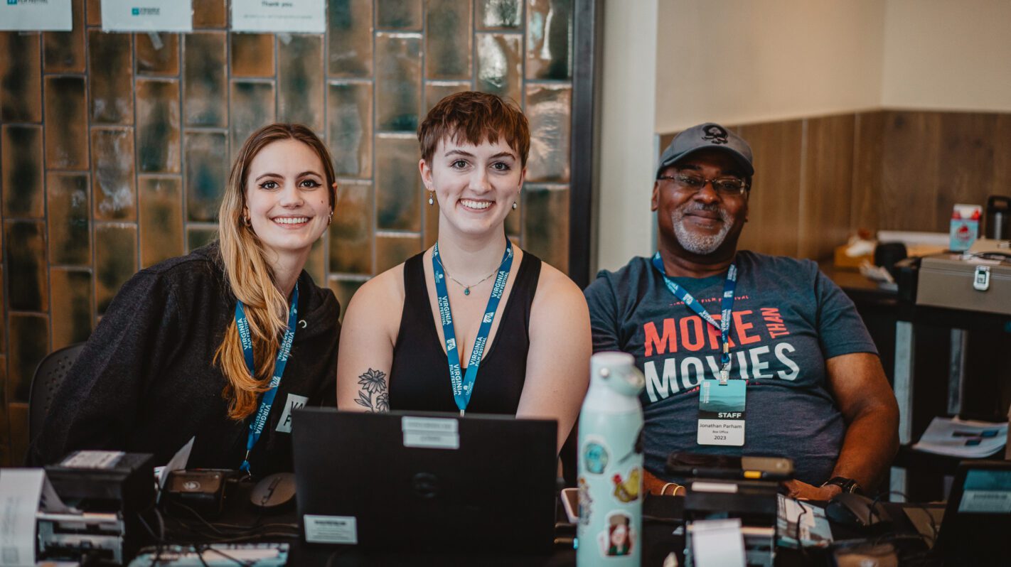 Three smiling film festival volunteers at a registration table. Staff badges visible.