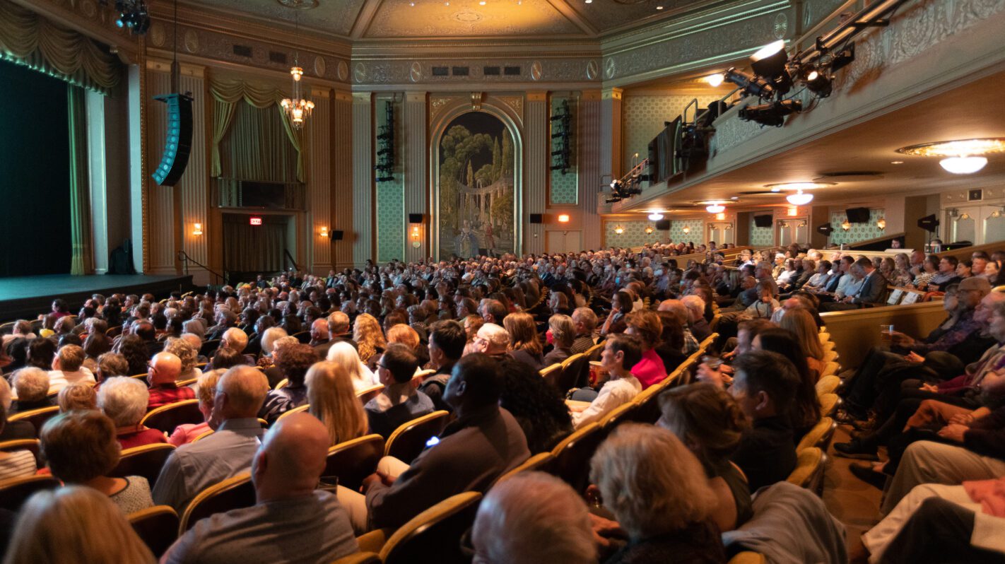 Audience seated in a grand theater, awaiting the performance.
