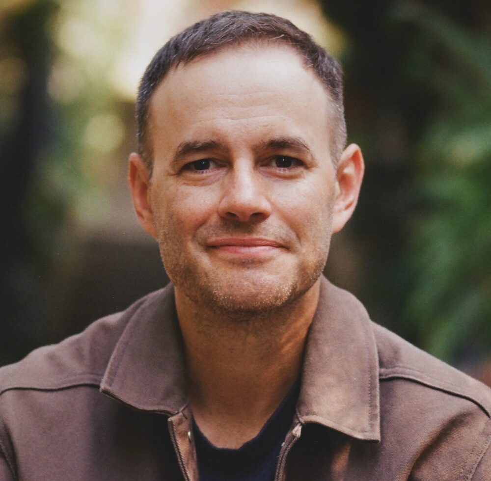 Clay Tweel portrait, wearing a brown jacket, outdoors.