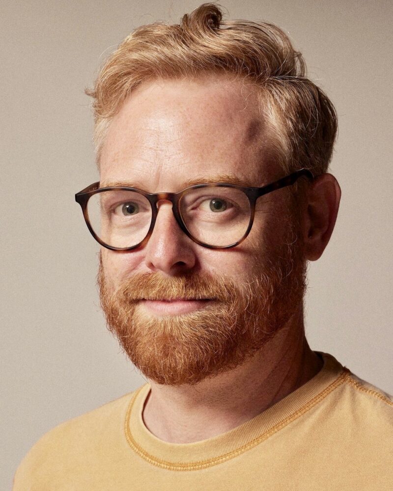Tyler Coates, a man with a red beard and glasses, smiles at the camera.