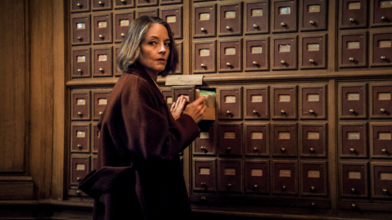 Woman with short gray hair looks through a card catalog in a library, possibly researching a private life.