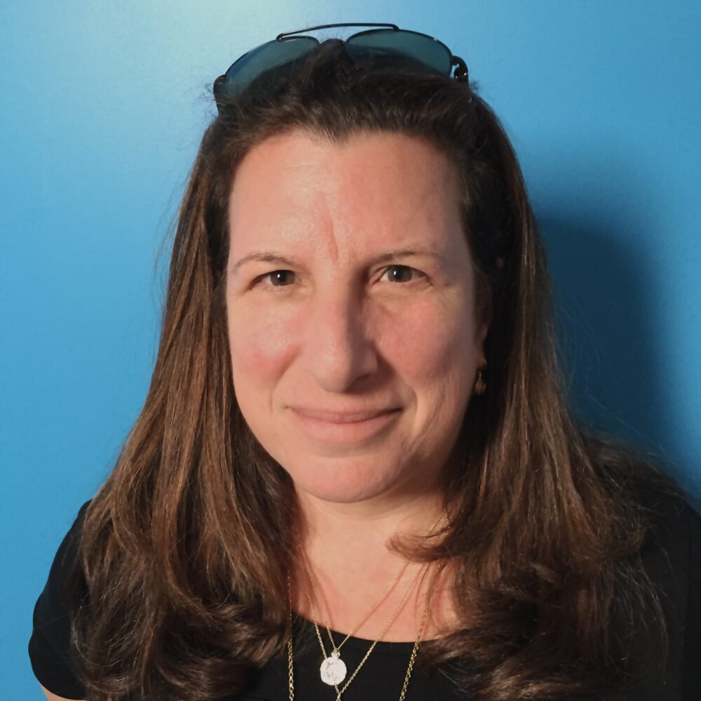 Alexa L. Fogel, headshot of a woman with sunglasses on her head, wearing gold necklaces.