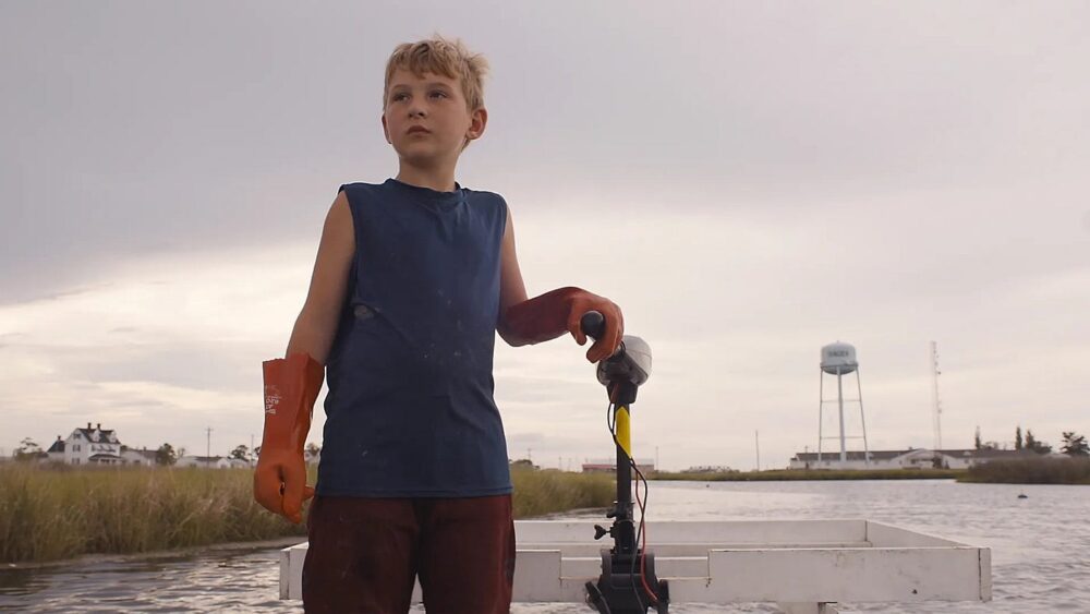 Young boy on a boat, wearing gloves, with a water tower in the background.