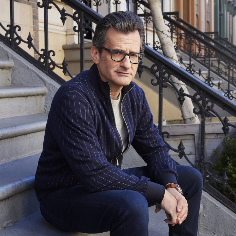 Ben Mankiewicz sitting on brownstone steps, wearing a blue jacket and glasses.