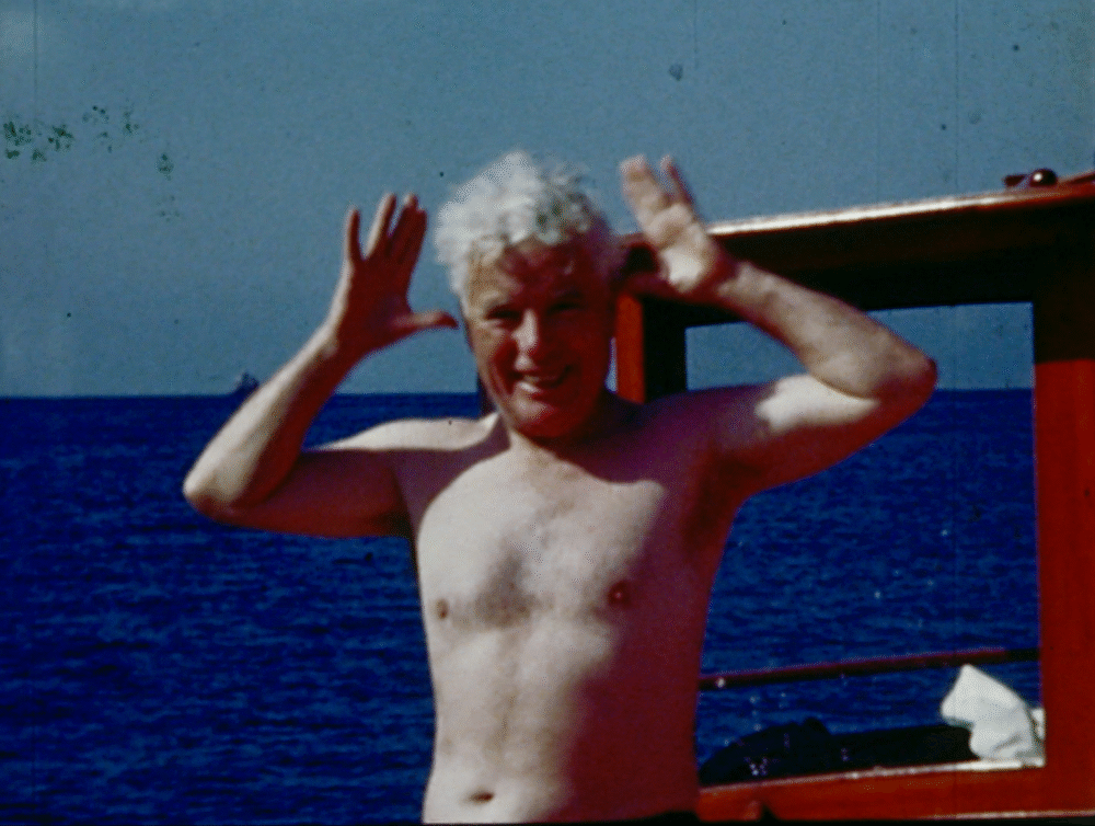 Man with white hair on a boat, making a playful gesture. Chaplin spirit.