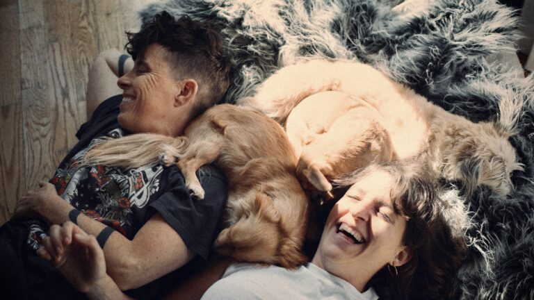 Two smiling women cuddling with dogs on a furry rug. Happy and relaxed indoors.