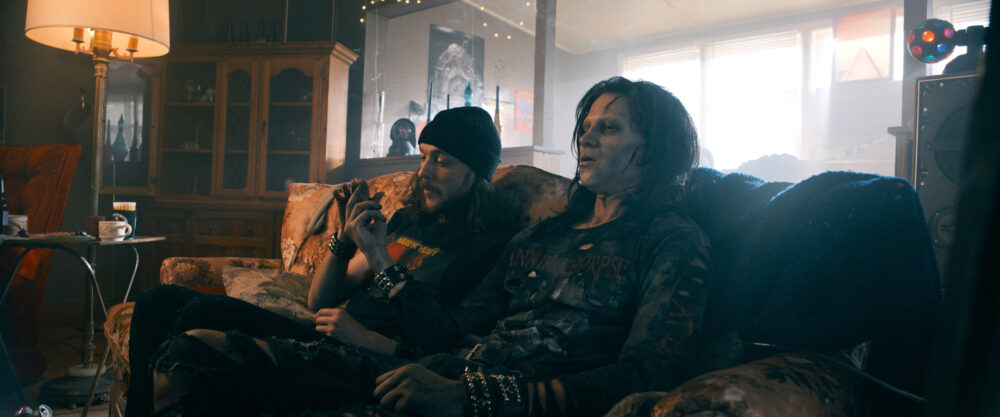 Two men on a couch, one in zombie makeup, possibly related to "Deathgasm II: Goremageddon.