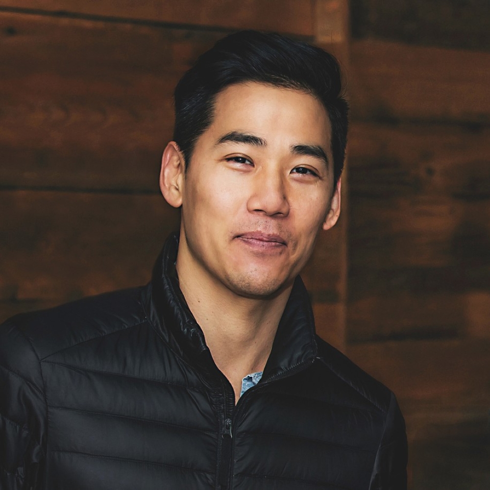 Portrait of David Usui in a black puffer jacket against a wood background.