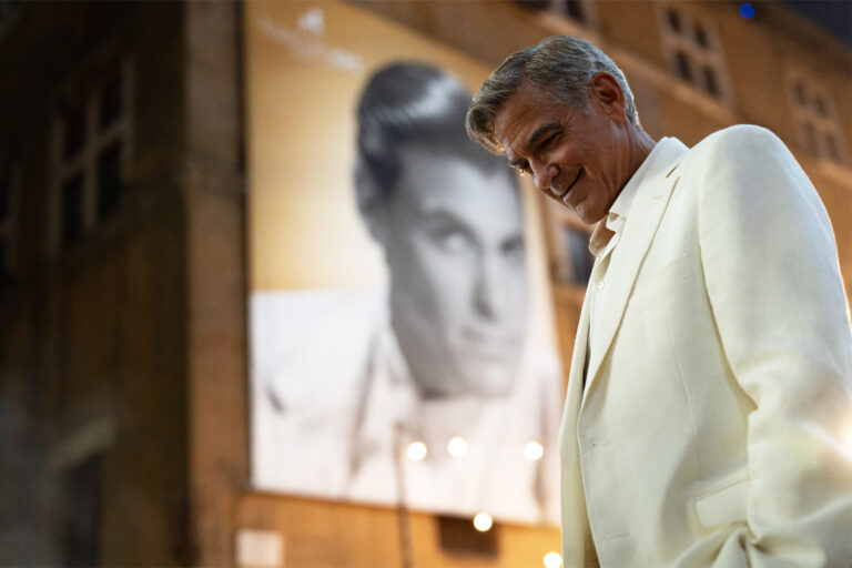 George Clooney in a white suit stands in front of a poster, smiling slightly.