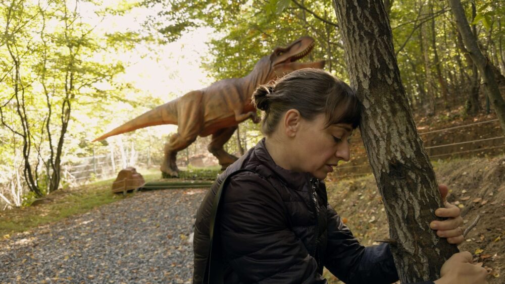 Woman hugs tree with T-Rex statue in background.
