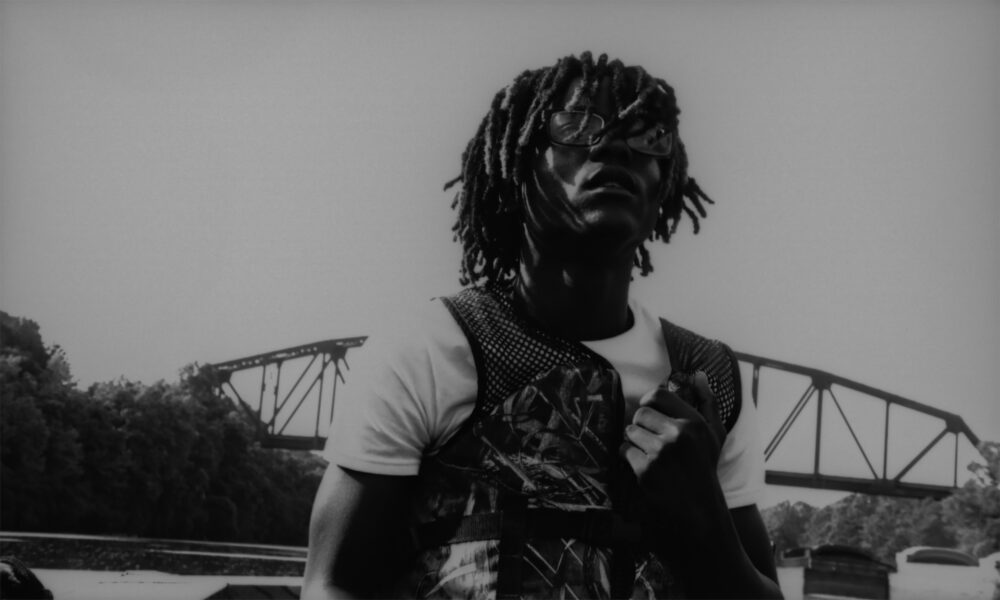 Black and white portrait of a man with dreadlocks and glasses in front of a bridge.