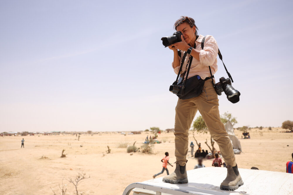 Photographer with cameras stands on vehicle in arid landscape, capturing images for Love+War.