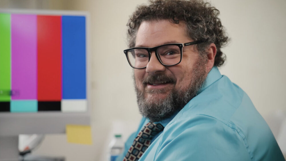Man with glasses smiling next to a monitor with a test pattern.