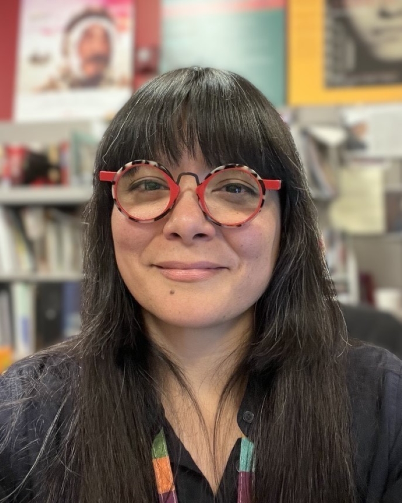 Melissa Bisagni, a woman with dark hair and round glasses, smiling in front of a bookshelf.