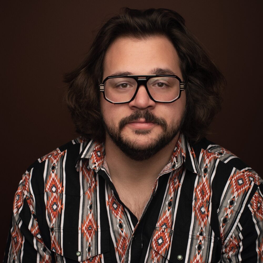 Michael Strassner in a patterned shirt and glasses against a brown background.