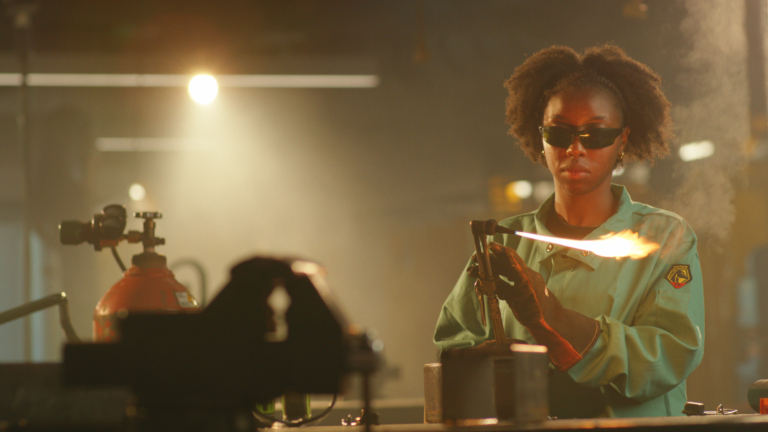 Woman welder using torch in industrial setting. Safety glasses, gloves, and green work shirt.