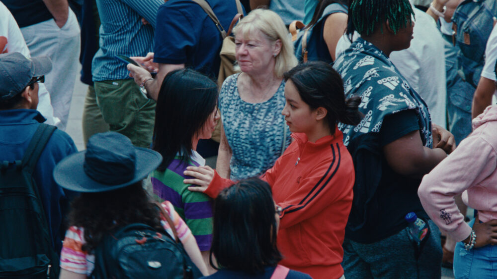 Crowd scene with two young women in foreground, one in red jacket, one in striped shirt, engaged in conversation.