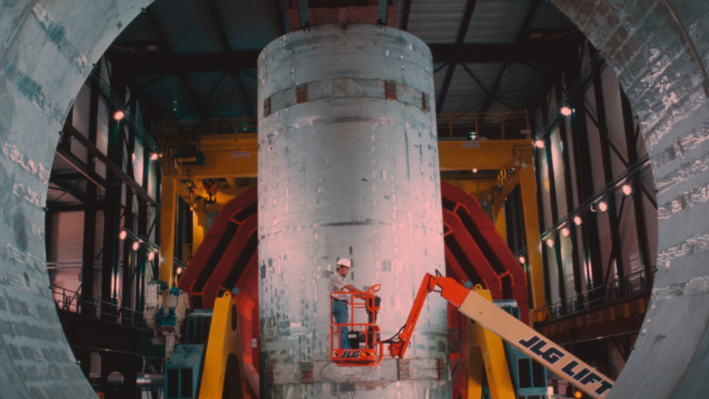 Worker inspecting a large rocket section inside a test facility with JLG lift, focusing on "The Peace Particle" concept.