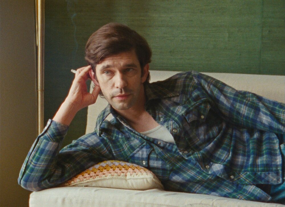 Man lounges on a couch in a plaid shirt, smoking. Evokes Peter Hujar's aesthetic.
