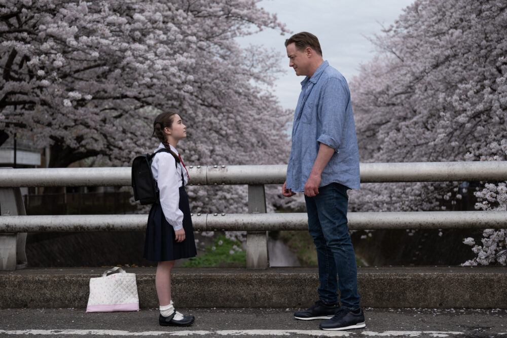 Man and girl in school uniform face each other under cherry blossoms.