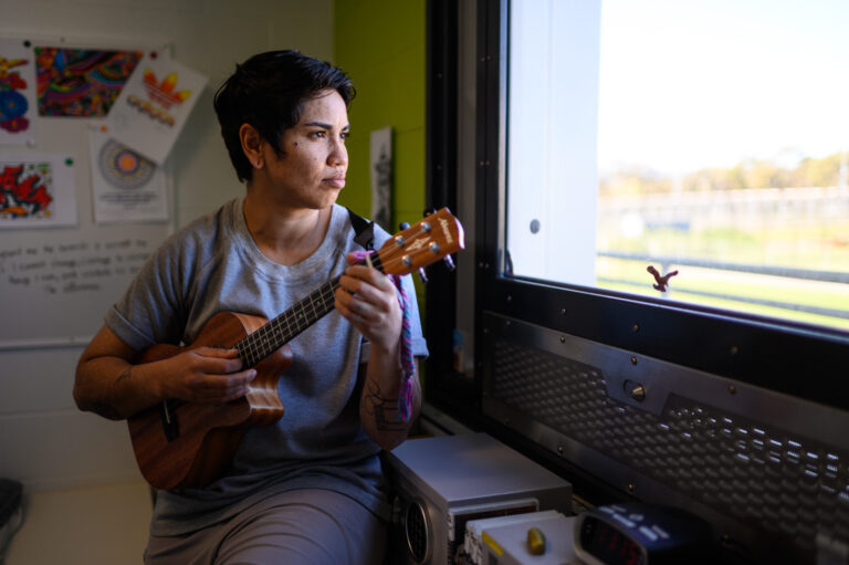 Woman in prison uniform plays ukulele by the window.