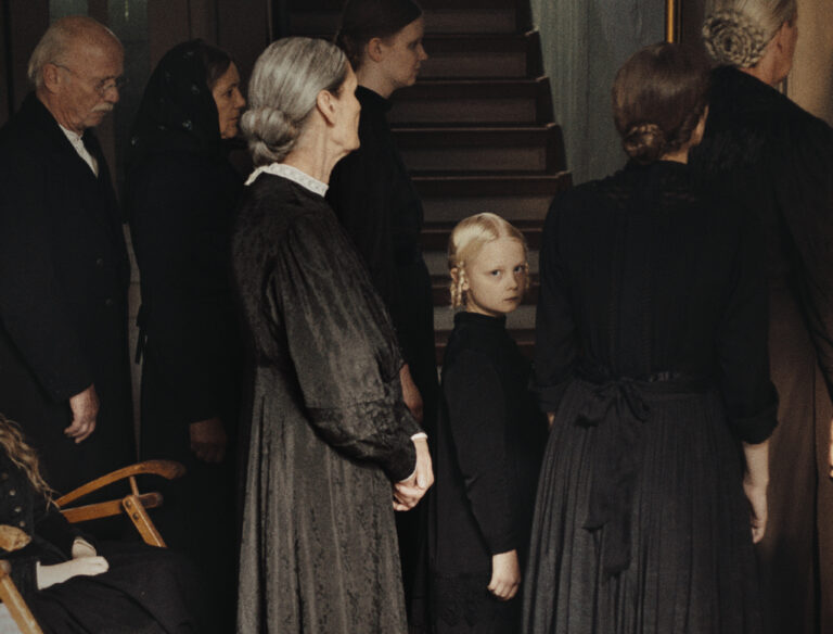 Mourners dressed in black at a somber funeral, a young girl looking at the viewer.