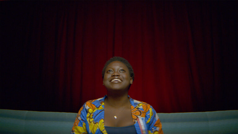 Woman in colorful jacket looking up against a red curtain. Speak.