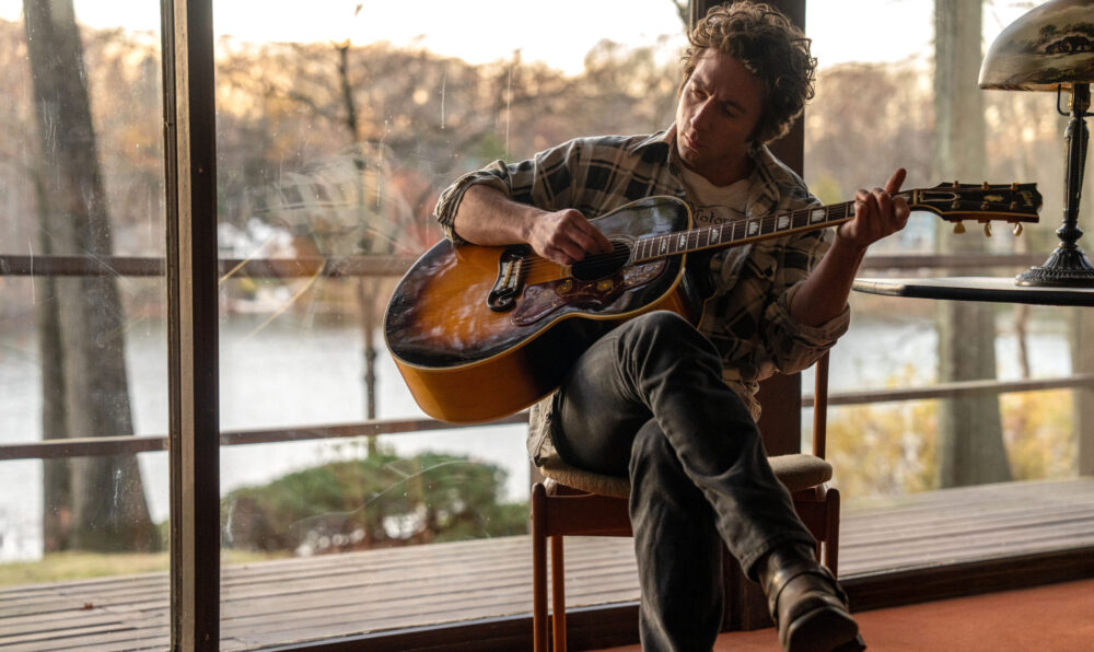 Man playing acoustic guitar indoors by a large window overlooking a lake.