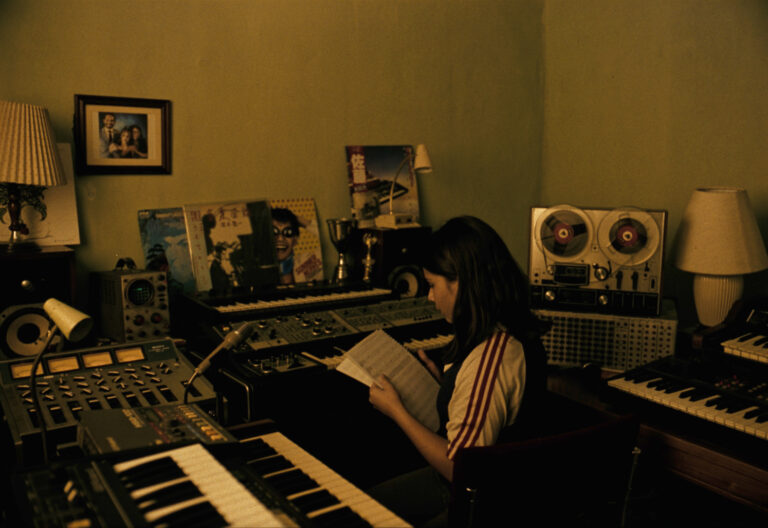 Woman in music studio with synths, reel-to-reel tape deck, and records, possibly working on "Synthesize Me" blog content.