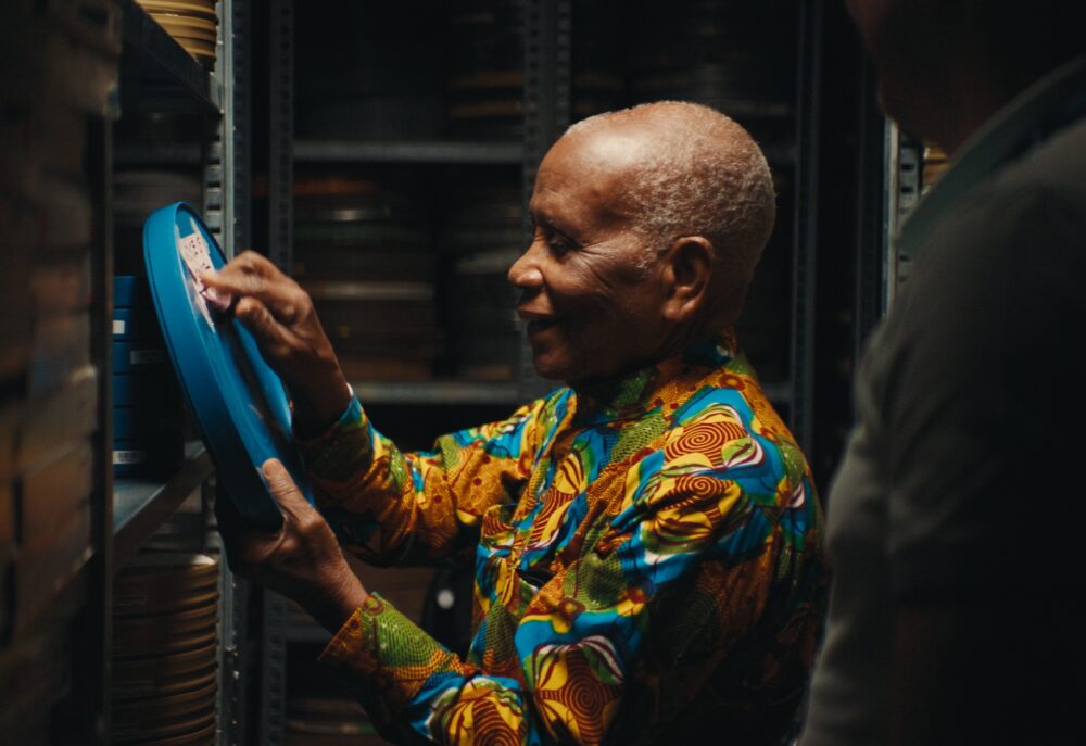 Man in colorful shirt inspecting film reel. "The Eyes Of Ghana" archival preservation.
