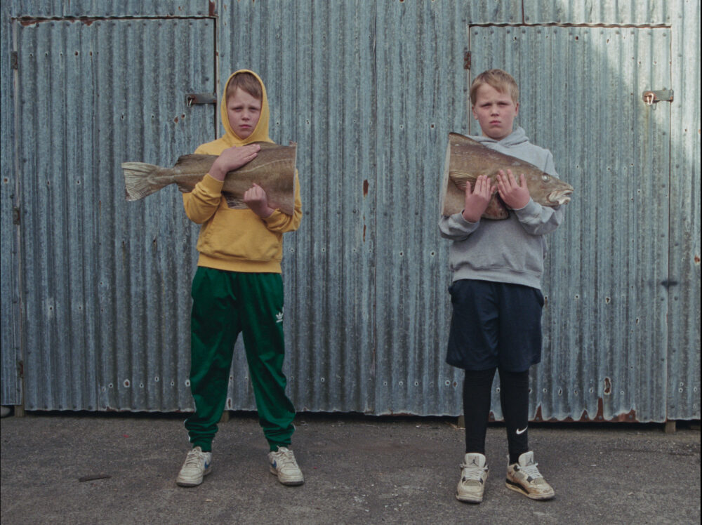 Two young boys holding dried fish against a corrugated metal wall.