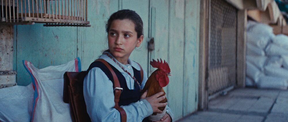 Young girl in school uniform holding a rooster, standing near a birdcage and sacks.