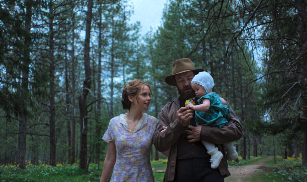 Family in "Train Dreams": Father, mother, and baby walk through a forest, the father holding a flower.
