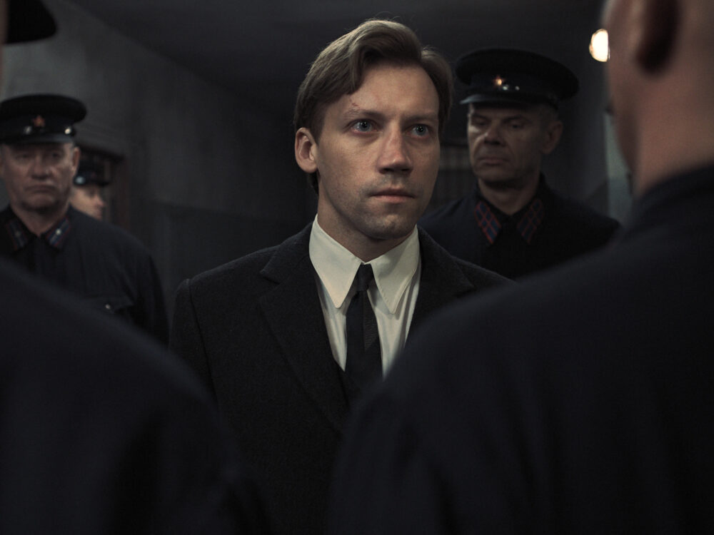 Prosecutor in suit surrounded by stern-faced men in military uniforms.