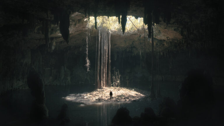 Person standing in a cenote with sunlight streaming through the opening above. Underland.