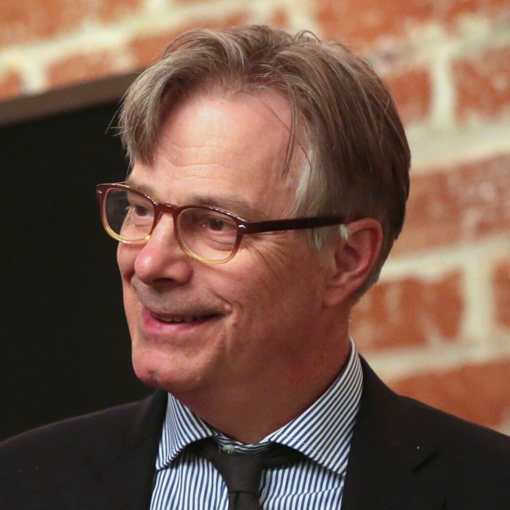 Whit Stillman smiles, wearing glasses and a striped shirt, against a brick wall.